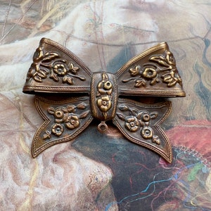 May include: A large, ornate, gold-toned metal bow with floral details. The bow has a loop at the top for hanging.