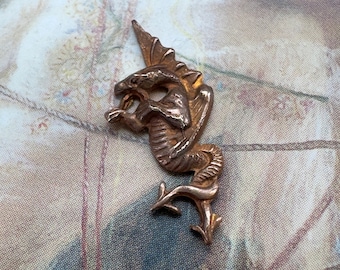 May include: A gold-toned metal dragon pendant with intricate details. The dragon is curled up with its wings spread out. The pendant is on a white background.