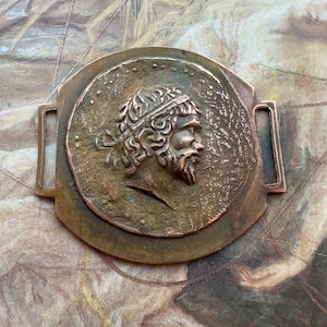 May include: A copper-toned metal belt buckle with a circular medallion featuring a profile of a man with a beard and a laurel wreath. The buckle has a rectangular frame with two rectangular loops.