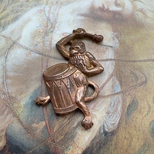 May include: A bronze-colored monkey figurine playing a drum. The monkey is holding a drumstick in its right hand and is beating the drum with its left hand. The drum is decorated with a pattern of lines.