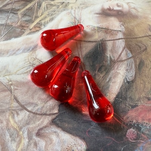May include: Five red glass teardrop-shaped ornaments with a pointed tip. The ornaments are arranged in a cluster on a textured surface.