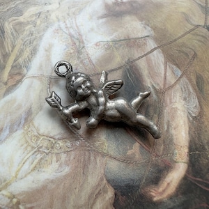 May include: A small, silver-toned charm in the shape of Cupid, holding a bow and arrow. The charm features detailed wings and a loop for attachment. The background is a vintage-style painting with soft, muted colors.