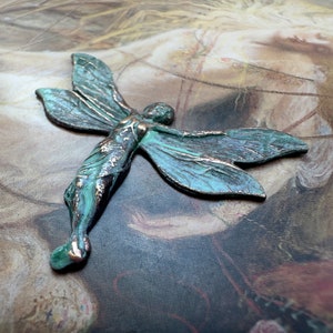 May include: A green and copper coloured metal fairy figurine with wings outstretched. The fairy is on a brown and white background.