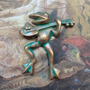 May include: A green and bronze frog figurine playing a guitar. The frog is standing on a textured surface with a painting of a woman in the background.