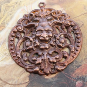 May include: A circular, bronze-toned metal pendant with a detailed face design. The pendant has an openwork pattern with a loop for hanging. The face has a stern expression, surrounded by swirling patterns and leaf motifs. The pendant is approximately 8 cm in diameter.