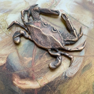 May include: A bronze crab figurine with detailed claws, legs, and body segments. The crab is positioned on a patterned background with muted colors. The figurine has a weathered appearance, suggesting an antique or vintage aesthetic.
