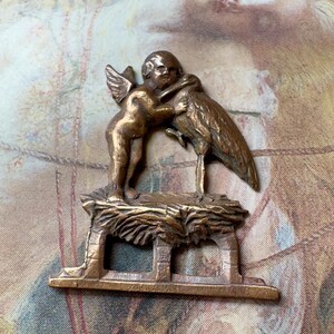 Puede incluir: Figura de bronce que representa un querubín alado abrazando una cigüeña. El querubín y la cigüeña están colocados sobre una base detallada y texturizada de forma rectangular. El diseño tiene una estética antigua.