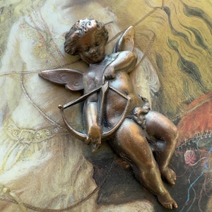 May include: A bronze-colored cherub figurine with wings and a bow and arrow. The cherub is posed in a seated position, with detailed facial features and curly hair. The background features a vintage-style painting with soft colors.