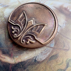 May include: A round, antique bronze button with an embossed floral design. The button features a raised, stylized floral motif in the center, surrounded by a circular border. The button's surface has a slightly aged, tarnished appearance, suggesting vintage craftsmanship.