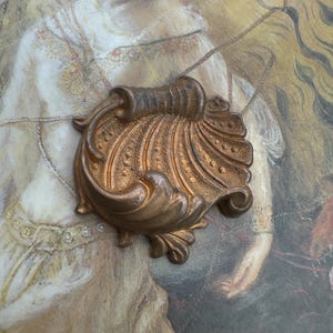May include: A bronze-colored, ornate decorative element with a shell-like design. The piece features intricate scrollwork and a textured surface, suggesting an antique or vintage aesthetic. The overall shape is symmetrical, with a central, ribbed section.