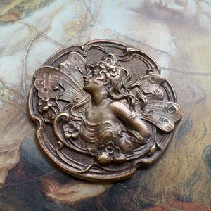 Puede incluir: Broche de estilo Art Nouveau en tono bronce que representa a una mujer con cabello suelto y alas de mariposa. El diseño detallado incluye detalles florales dentro de un marco circular decorativo. La broche tiene una estética vintage.