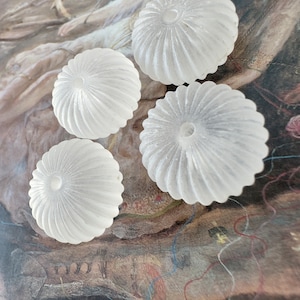 Könnte beinhalten: Vier weiße, gefrostete Glasperlen mit einem geriffelten Design. Die Perlen haben ein Loch in der Mitte und sind in einer Gruppe auf einer strukturierten Oberfläche angeordnet.