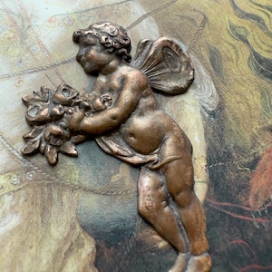 May include: A bronze-colored cherub figurine holding a bouquet of flowers. The cherub has detailed wings and is in a side-facing pose. The figurine is likely a decorative element, possibly for crafts or home decor.