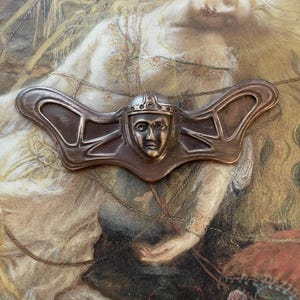 May include: A bronze-toned metal brooch featuring a central face wearing a helmet and crown, flanked by stylized wings. The brooch is set against a backdrop of a vintage painting with a classical theme.
