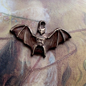 May include: A bronze-colored bat pendant with outstretched wings. The bat has detailed features, including a textured body and a loop at the top for hanging. The pendant is likely for jewelry or crafting.