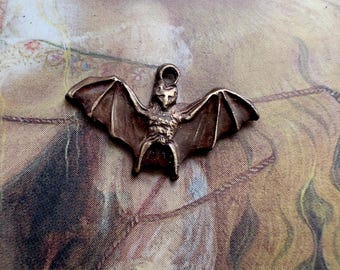 May include: A bronze-colored bat pendant with outstretched wings. The bat has detailed features, including a textured body and a loop at the top for hanging. The pendant is likely for jewelry or crafting.