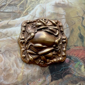 May include: A vintage brass brooch with an ornate design featuring a central oval shape surrounded by stylized floral motifs.