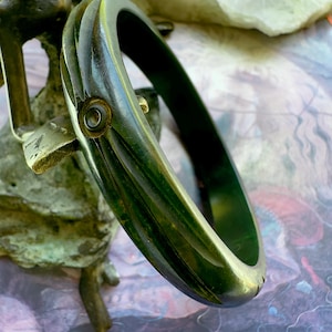 May include: A dark green, polished bangle bracelet featuring a carved, ribbed texture along its outer surface and a small, circular inlaid detail. The bangle has a smooth, rounded interior and a glossy finish that reflects light across its curved form.