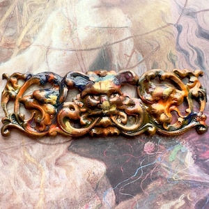 May include: A gold-toned metal decorative piece with a detailed, ornate design. The piece features a central grotesque face with swirling, floral patterns on either side. The piece is painted with a multi-colored, iridescent finish.