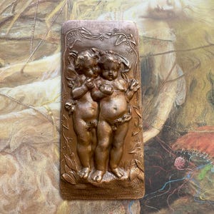 May include: A rectangular, bronze-colored relief sculpture featuring two cherubic figures embracing. The figures are depicted with wings and are surrounded by floral and foliate details. The relief has a detailed, antique appearance.