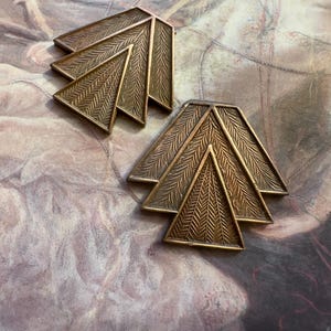 May include: Two bronze-colored, geometric brooches. Each brooch features a triangular design with a textured, leaf-like pattern. The brooches are arranged on a patterned background, showcasing their intricate details and metallic finish.