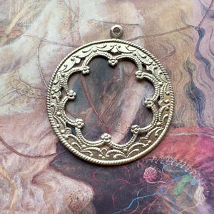 May include: A silver-toned metal pendant with an intricate, openwork design. The pendant is oval-shaped and features a floral motif.