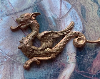 Op de afbeelding: Een gouden metalen drakenbeeldje met vleugels en een gekrulde staart. De draak is gedetailleerd met schubben en heeft een woeste uitdrukking.