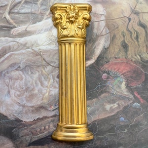 Puede incluir: Una columna decorativa de color dorado con un fuste estriado y un capitel ornamentado. La columna está hecha de un material liso y brillante y tiene un diseño clásico.