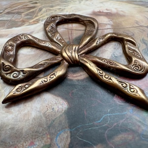 May include: A gold-toned metal bow with intricate swirling designs. The bow is a decorative piece with a vintage aesthetic.
