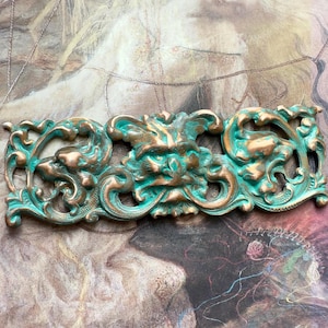 May include: A decorative metal ornament with a green patina finish. The ornament features a stylized face with intricate floral details.