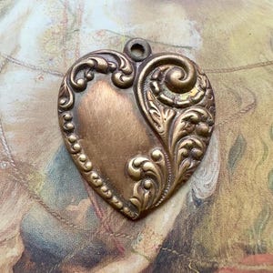 May include: A vintage heart-shaped pendant with an ornate design. The pendant is made of a bronze-colored metal and features a blank oval space on the left side. The right side is decorated with swirling floral patterns. A small loop is at the top for hanging.