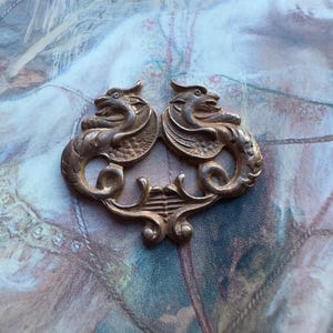 May include: A gold-toned metal decorative piece featuring two stylized dragons facing each other. The dragons have intricate details and are connected by a decorative scrollwork design.