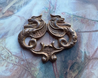 Op de afbeelding: Een decoratief stuk van verguld metaal met twee gestileerde draken die elkaar aankijken. De draken hebben ingewikkelde details en zijn verbonden door een decoratief rankwerk.