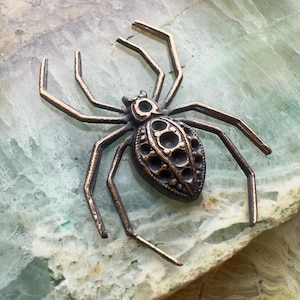 May include: A bronze-toned spider brooch with intricate detailing. The spider's body features a textured design with multiple circular cutouts. The legs are thin and angular, extending outwards. The brooch is set against a light green and white stone surface.
