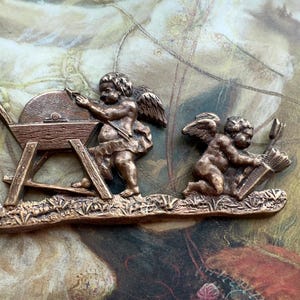 May include: A bronze-colored decorative relief sculpture featuring two cherubs. One cherub sharpens a tool on a grindstone, while the other kneels, holding a quiver of arrows and a bow. The sculpture is set against a muted, textured background.