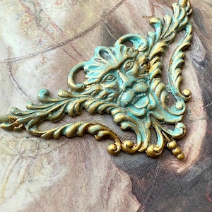 May include: A decorative metal ornament with a green and gold finish. The ornament features a stylized face with a beard and a floral design.