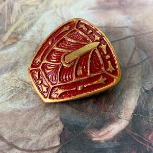 May include: A red and gold shield-shaped brooch with intricate details. The shield has a gold design of a sword and other decorative elements.