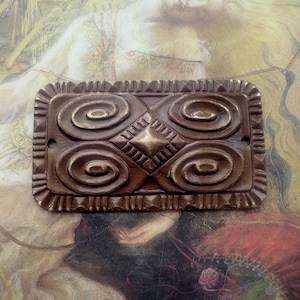 May include: A rectangular, bronze-colored decorative plaque with a detailed, carved design. The plaque features four swirling spiral patterns and a central diamond shape. The edges are framed with a textured border. The background is a painting.