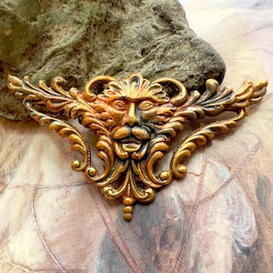 May include: A gold-toned metal brooch with a detailed design of a grotesque face with a leafy background.