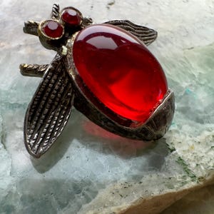 Könnte beinhalten: Silberfarbene Insektenbrosche mit einem großen, ovalen, roten Edelstein als Körper. Zwei kleinere rote Edelsteine ​​sind am Kopf angebracht. Die Flügel und Beine sind mit geätzten Linien detailliert, was eine Vintage-Ästhetik erzeugt.