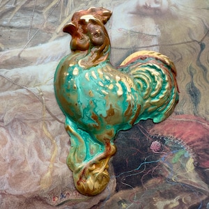 以下が含まれることがあります： ターコイズ、ゴールド、ブロンズ仕上げの装飾的な雄鶏の彫刻。 セラミック製の雄鶏は質感のある表面を持ち、ヴィンテージ絵画の背景に配置されています。