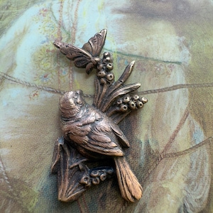 May include: A bronze metal brooch depicting a bird on a branch with a butterfly. The bird and butterfly have detailed features. The brooch is set against a soft, patterned background. The overall design has an antique aesthetic.