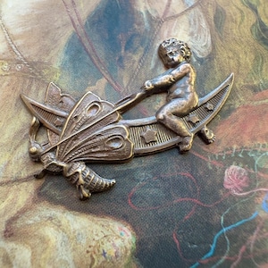 May include: A bronze-toned metal brooch featuring a cherub riding a crescent moon and pulling a butterfly. The butterfly has detailed wing patterns. The cherub is sculpted with fine details. The brooch has an antique or vintage aesthetic.
