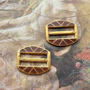 Puede incluir: Dos hebillas de metal dorado con un patrón geométrico marrón y rojo. Las hebillas tienen dos barras horizontales.