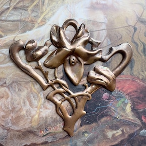 May include: A bronze-colored, Art Nouveau-style decorative element featuring a stylized flower with curved petals and leaves. The design includes intricate, flowing lines and organic shapes, suggesting a botanical theme. The piece is set against a backdrop of a vintage illustration.