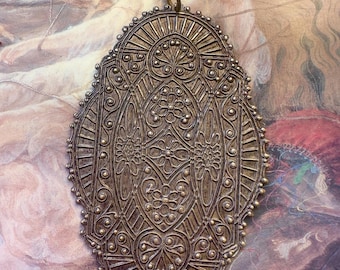 Puede incluir: Un gran colgante ovalado de latón con intrincados diseños en relieve detallados. El colgante tiene un bucle en la parte superior para colgar.