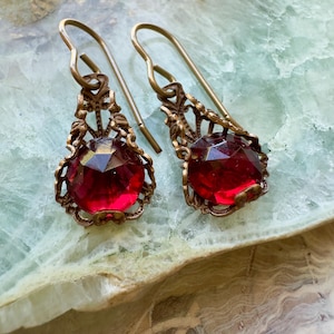 May include: A pair of vintage-style earrings featuring large, faceted, ruby-red glass stones. The stones are set in ornate, filigree-style brass settings with long, curved ear wires. The earrings are displayed on a light green and white surface.