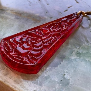 May include: A red, pendant-shaped charm with a carved rose design. The charm has a gold-colored clasp at the top. The pendant is made of a translucent, glossy material.