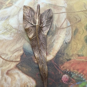Könnte beinhalten: Ein bronzefarbener, blattförmiger Anhänger mit detaillierten Adern. Der Anhänger hat eine strukturierte Oberfläche und eine kleine Schlaufe zum Aufhängen. Das Blattdesign ist ein von der Natur inspiriertes Accessoire.