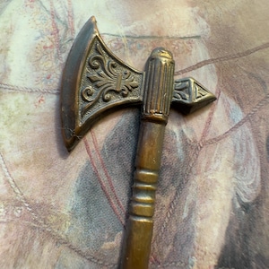 May include: A miniature bronze axe with an ornate, engraved blade and a wooden handle. The axe head features a detailed floral design. The handle is a dark brown color. The axe is a decorative item.